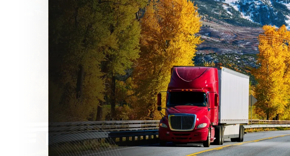 A red semi-truck with a white trailer drives along a highway lined with golden autumn trees and snow-capped mountains.