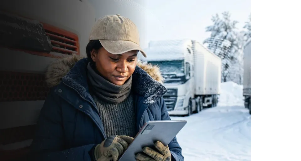A person in a winter parka and cap uses a tablet in a snowy parking lot with semi-trucks in the background.